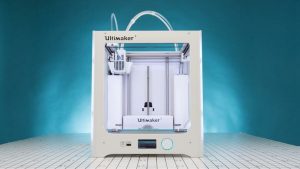 An Ultimaker 3 sits on a white counter in front of a blue background. The Ultimaker is a large white cube with a glass front so that you can watch as objects print.