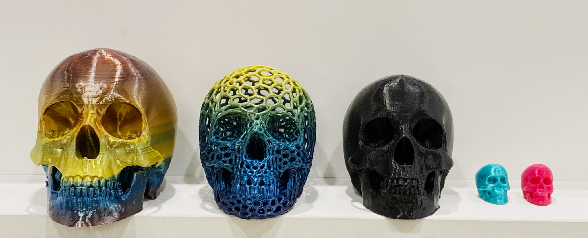 5 3-D printed skulls, from largest to smallest. The first two are using rainbow silk PLA filament, the third using black PLA filament, and the last two tiny skulls using silk teal and hot pink PLA filaments.
