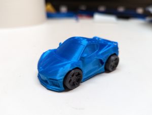 A 3-d printed miniature race car in silk blue with black tires sits on a white surface.