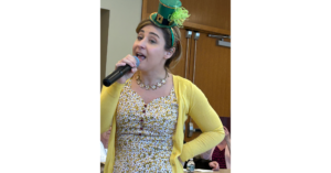 A woman with a microphone entertaining memory cafe guests on 3-7-2025 while wearing a leprechaun hat.