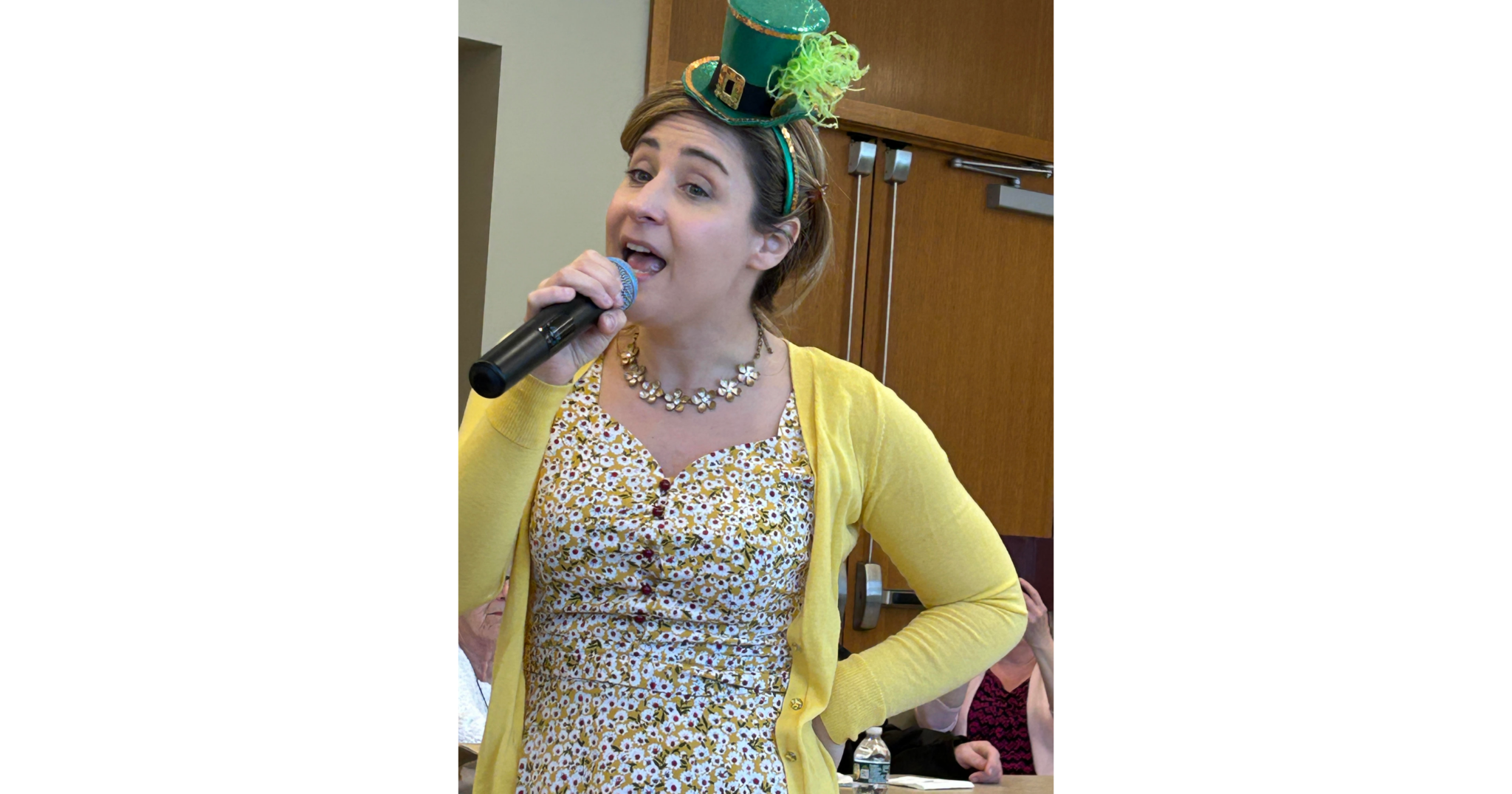 A woman with a microphone entertaining memory cafe guests on 3-7-2025 while wearing a leprechaun hat.